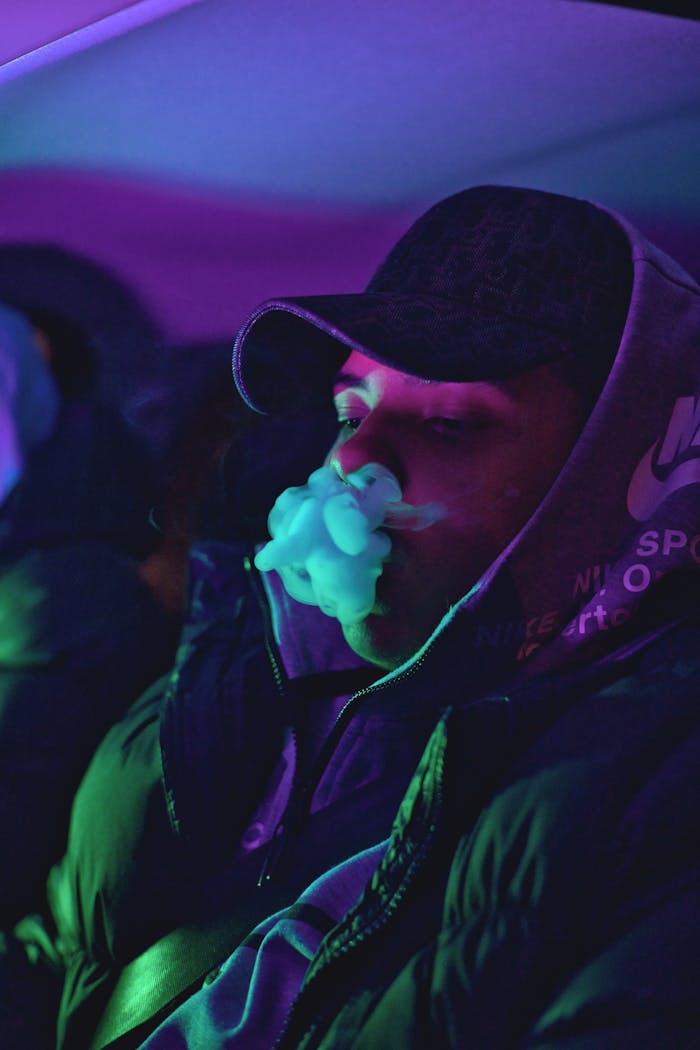 Man in a cap and hoodie exhaling vapor in a neon-lit setting, creating a moody and modern vibe.