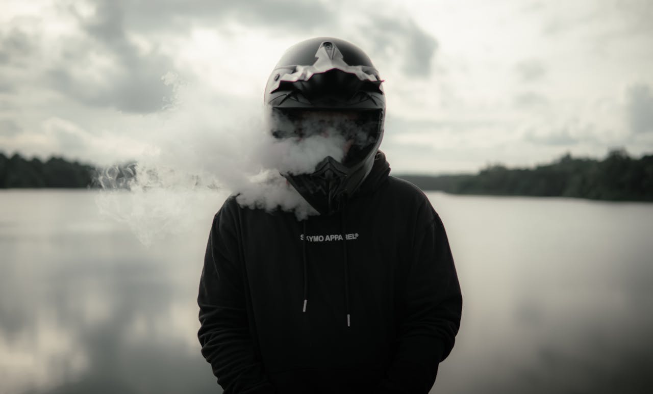 A motorcyclist stands by a tranquil lake, exhaling vapor, with a cloudy sky backdrop.